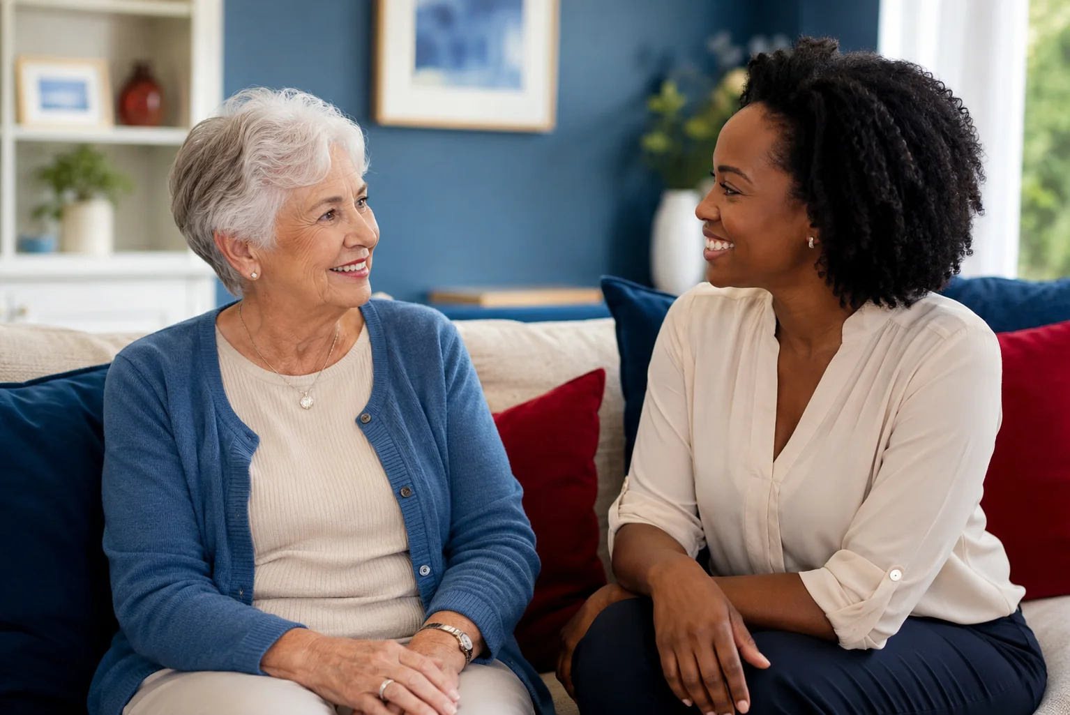 Senior Caucasian woman and African American caregiver sitting on a sofa and smiling during a warm conversation at home.
